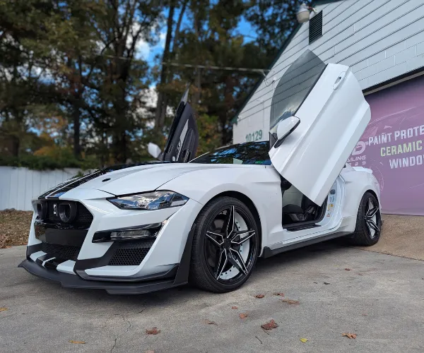 A front three-quarter view of a black &white sports car parked inside a brightly lit detailing garage, showcasing a high-gloss, reflective ceramic coating finish on the hood and body.